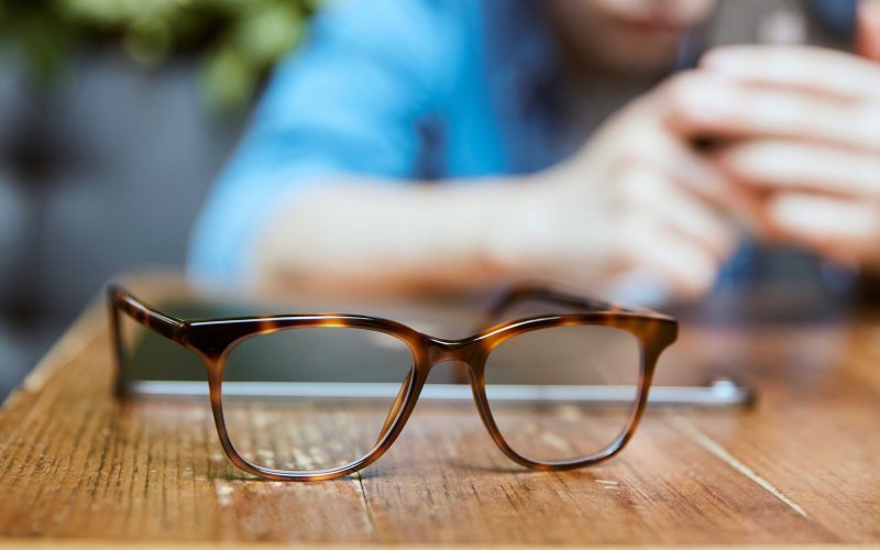 A pair of tortoise glasses on a wooden table with a man on a phone in the background