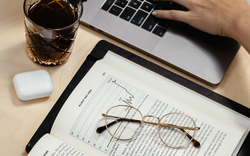 Glasses on a book next to someone typing on a laptop with a cup of coffee